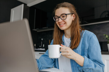 Relaxed female freelancer relaxing sitting with cup of coffee in front of laptop, working or studying remotely from home, drinking from mug taking a break