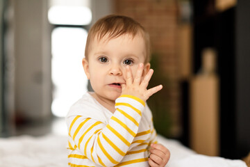 Teething concept. Portrait of little baby girl sitting on white bedsheets and biting finger in...