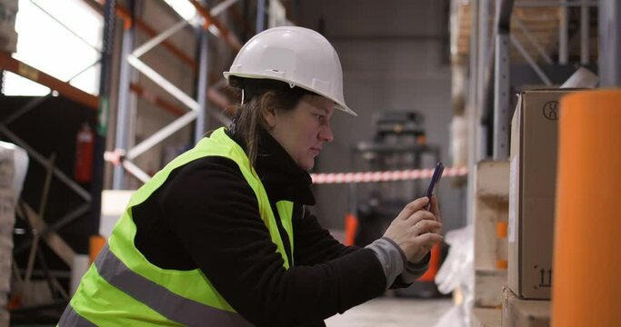 4k slow-motion video of the female warehouse manager using the mobile phone to scan the boxes while sorting the storage.  The coworkers can be seen in the background doing their work.