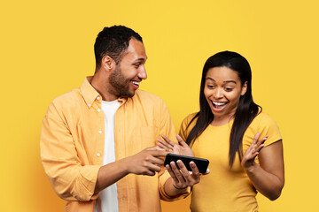 Glad young african american guy showing phone to shocked wife, isolated on yellow background, studio