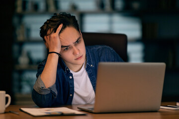 Hopeless frustrated man looking at laptop screen, dark office interior