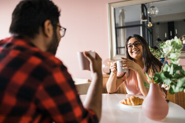 Happy couple eating breakfast and talking at dining table in morning. Indian girl and latin guy. Relationship and diversity concept