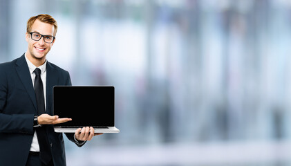 Business man in glasses spectacles, black suit showing laptop with empty mockup screen, blurred interior background, with copy space. It expert,  technician repair service, tech support, maintenance.