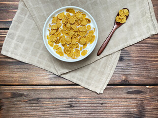 Corn flakes with milk in white plate and wooden spoon on beige towel. Wooden background, top view.