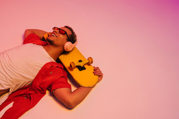 Cheerful african guy laying with skateboard isolated over gradient neon studio background