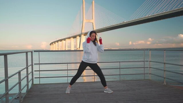Woman jump in leg kickboxing kick hands wrapped in red boxing tapes with bridge and blue sky background. Middle age female athleteboxing fighter training outside dressed hoodie. Full length 