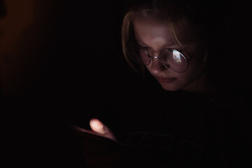 a teenage girl of European appearance in glasses for vision, sits in a dark room looking at the phone, the light falls on the girl's face the girl is sitting sad on the phone bullying on the Internet,