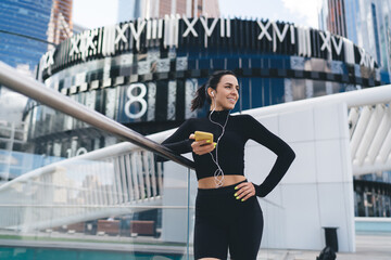 Cheerful woman standing with smartphone and listening to music in earphones