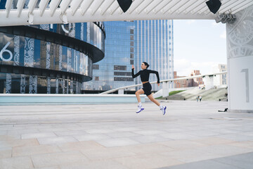 Focused sportive woman running along urban buildings