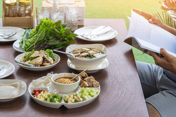 Spicy Vietnamese Pork Sausage Salad (Yum Moo Yor). Pork sausage spicy salad Thai and Vietnam food with other appetizing dishes and vegetables on wooden table with young man read a book before eating.