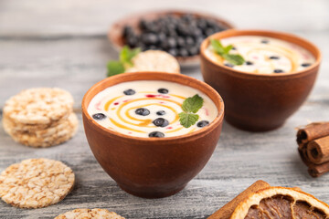 Yoghurt with bilberry and caramel in clay bowl on gray wooden, side view, selective focus.