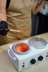 Hot orange wax used for hair removal. Close-up of depilation master hands in black protective gloves holds spatula with melted wax. Device for melting wax. Vertical Screen Orientation Video 9:16