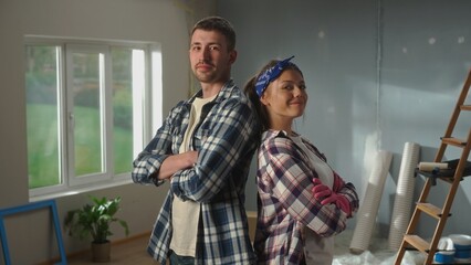 Portrait of woman in pink rubber gloves and man is crossing their arms, looking seriously at camera and smiling. Young couple in plaid shirts is posing against backdrop of room in process of being - Powered by Adobe