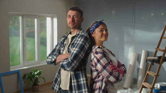 Portrait of woman in pink rubber gloves and man is crossing their arms, looking seriously at camera and smiling. Young couple in plaid shirts is posing against backdrop of room in process of being