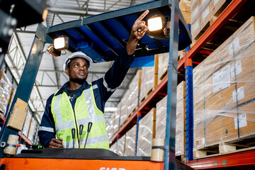 Fototapeta na wymiar Handsome Professional Black Worker Wearing Safety Vest and Hard Hat Charmingly Smiling and Looks Into the Distance. Background Big Warehouse with Shelves full of Delivery Goods.
