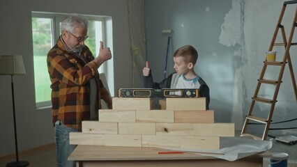 Grandpa shows his grandson how the water level installed on a brick wall works. Elderly man and young guy show thumbs up. The concept of family, joint renovation in the apartment.