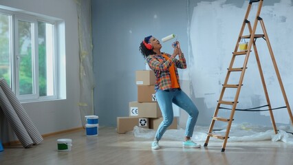 Young african american woman in big wireless red headphones is enjoying music. Black female in checkered shirt sings into paint roller as if into microphone and dances. Ladder, cardboard boxes, window
