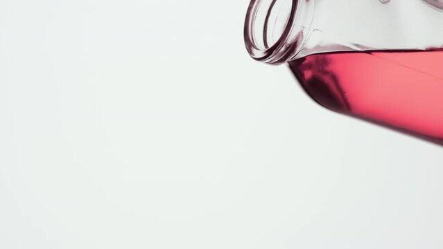 Side view macro shot of reagent bottle is being tilted and pink fluid pours from it on grey background | Abstract face care cosmetics ingredients formulating concept