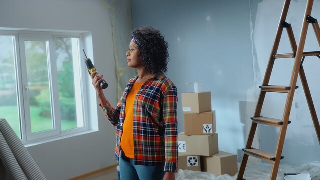 Young African American woman is turning on an electric screwdriver. Black female standing against the background of cardboard boxes, ladder and window and looking at drill.