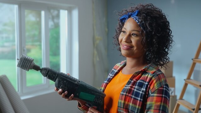 A Young African American Woman Holds An Electric Hammer Drill With A Nozzle For Drilling Holes In Her Hands. A Black Woman Poses In Front Of A Window And Smiles. The Concept Of Repair In The Apartment