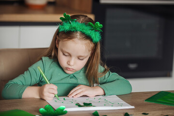 A little girl with a shamrock headband draws and cuts green shamrocks for St. Patrick's Day at her...
