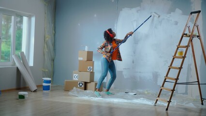 African American woman in red headphones paints wall with white paint using long paint roller and enjoy the music. Black female making repairs in an apartment. Window, ladder, cardboard boxes.