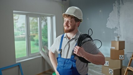 Foreman or electrician in blue overalls, white helmet and coil of black cable in hands. Redhead man...