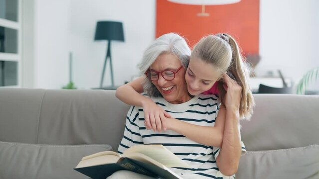 Video of kind senior woman reading a book while her granddaughter hugs her from behind in the sofa at home.