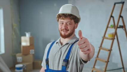 Repairman worker in blue overalls and white helmet is smiling and showing thumbs up. Portrait of redhead man is posing against backdrop of apartment in process of renovation.