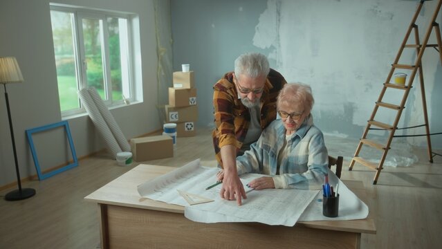 Elderly Man And Woman Are Looking Through Sheet With Plan Of An Apartment And Discussing Renovation Project. Aged Couple Is Sitting At A Table And Planning The Improvement Of Their Home.