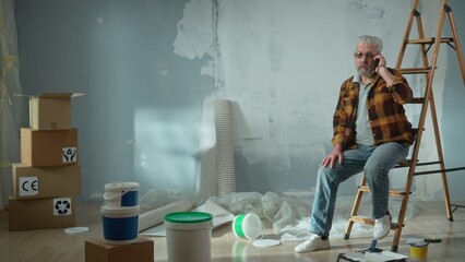 Tired elderly man is sitting on a ladder and calling a master to repair an apartment using a mobile...