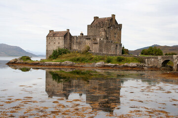 Castillo  de Eilean Donan, Escocia, y su reflejo en el agua del lago Duich, rodeado de verde y con el agua llena de algas en un d&iacute;a gris