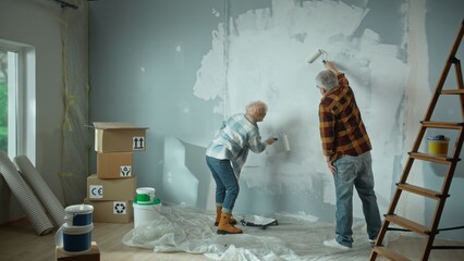 Elderly man and woman are painting wall with white paint using paint rollers. Couple of pensioners are making repairs to their apartment, in the background of window, stepladder, cardboard boxes. Back
