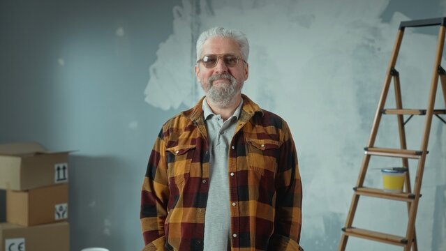 Elderly gray haired man with beard in glasses is looking at camera and smiling. Portrait of pensioner in plaid shirt is posing in an apartment against the background of a wall painted with white paint