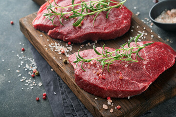 Raw beef steak. Marbled raw fresh Ribeye steak with rosemary, salt and pepper on cutting board on dark concrete background. Raw beef steak and spices for cooking.