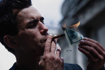 Young brutal businessman lighting cigar with $100 dollar bill. Horizontal image.