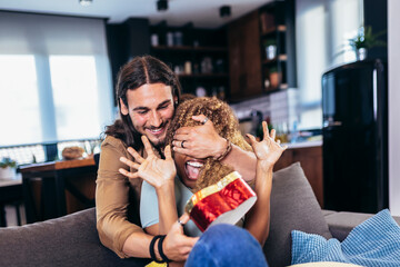 Beautiful young couple on couch at home. Handsome guy giving a gift box to his girlfriend.