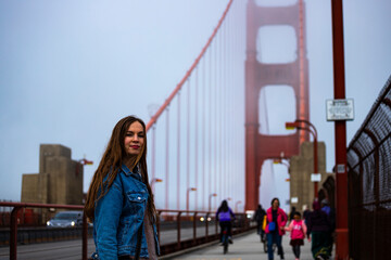 a beautiful long-haired model in a denim jacket stands on the famous golden gate bridge in san...