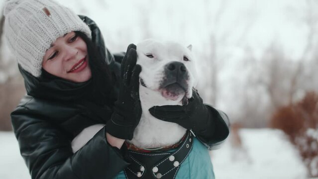 Young caucasian woman in winter clothes pats a white pit bull's face while squatting down afternoon in winter outdoor close up. Woman enjoys playing with her dog