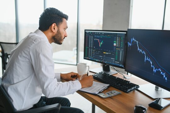 Young indian business man trader looking at computer screen with trading charts reflecting in eyeglasses watching stock trading market financial data growth concept, close up.