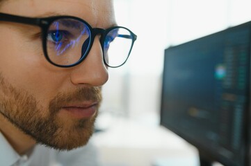 Young business man trader wearing glasses looking at computer screen with trading charts reflecting in eyeglasses watching stock trading market financial data growth concept, close up.