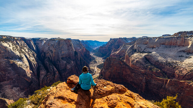 Girl in a blue jacket sits on rocks and admires the unique canyon from Observation Point (East Mesa Trail) in Zion National Park, Utah, USA. Winter in Utah