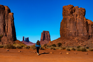 girl in denim jacket walks through monument valley with massive monuments in the background; walk...