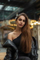 Fototapeta na wymiar Beautiful young woman in black stylish clothes with a one-shoulder top in a fashionable down jacket sits on a bench and poses in the city. Street, beauty and fashion