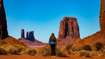 girl in denim jacket walks through monument valley with massive monuments in the background; walk...