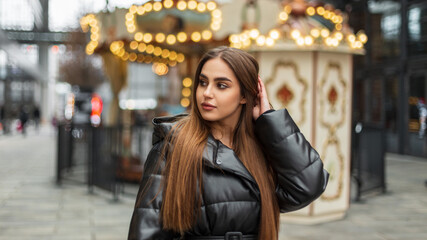 Fashionable beautiful elegant woman model with brunette hair in a stylish black down jacket walks in the city near a vintage carousel with lights. Urban winter female portrait of pretty girl