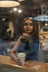 Fashionable beautiful elegant woman in a fashion sexy black one-shoulder top sits in a cafe near the window with reflection and drinks coffee