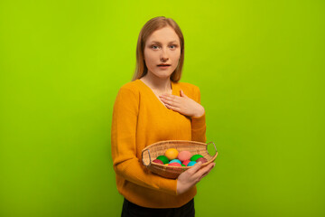 Open eyed surprised shocked young woman holding wicker basket with colorful painted dyed easter eggs, wearing yellow sweater and looking at camera isolated on green background.

Easter Day concept.
