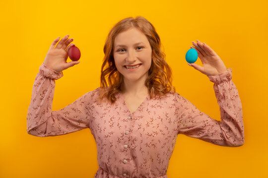 Happy smiling young woman with curly wavy hair holding red and blue painted easter eggs or two dyed easter eggs, wearing spring floral pink dress isolated on yellow background.

Easter Day concept.