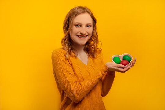 Happy smiling cheerful girl holding heart shaped plate saucer with three colorful painted dyed easter eggs, wearing yellow sweater isolated on yellow background.

Easter Day celebration concept.